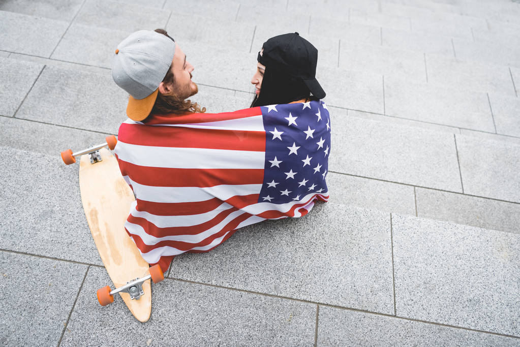 високий кут зору пари з американським прапором на плечах, що сидять на сходах біля скейтборду, дивлячись один на одного
 - Фото, зображення
