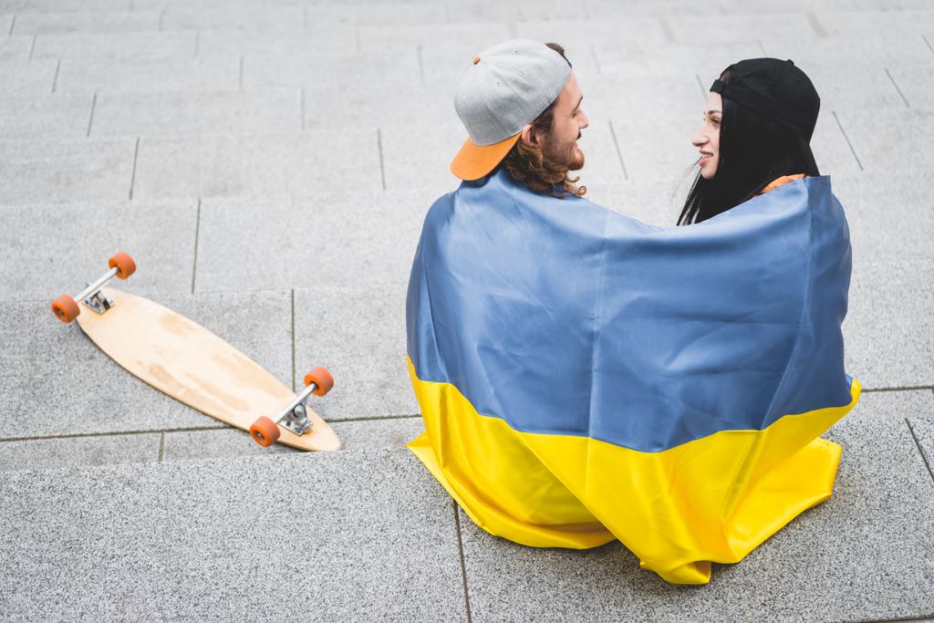 high angle view of couple with ukrainian flag sitting on stairs near skateboard, looking at each other - Photo, Image