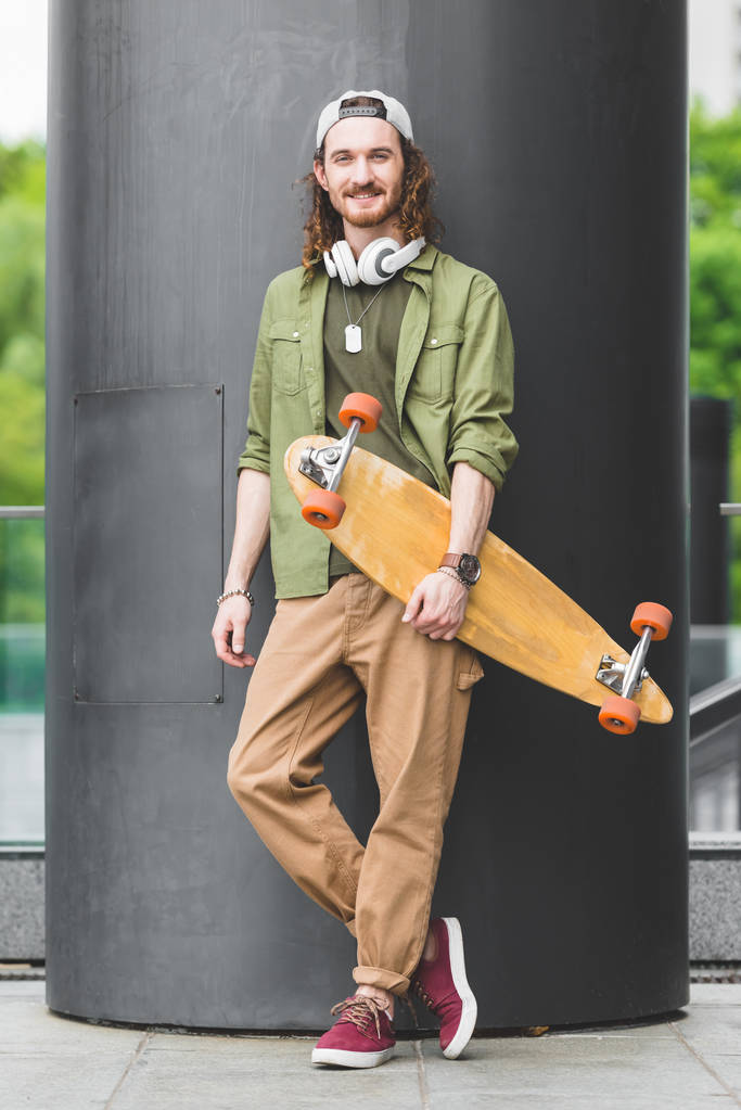 pohledný muž, který drží skateboard v ruce, stojí na střeše a dívá se na kameru - Fotografie, Obrázek