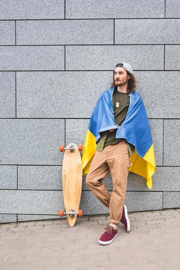 calm man in casual wear and ukrainian flag standing near wall with skateboard - Photo, Image