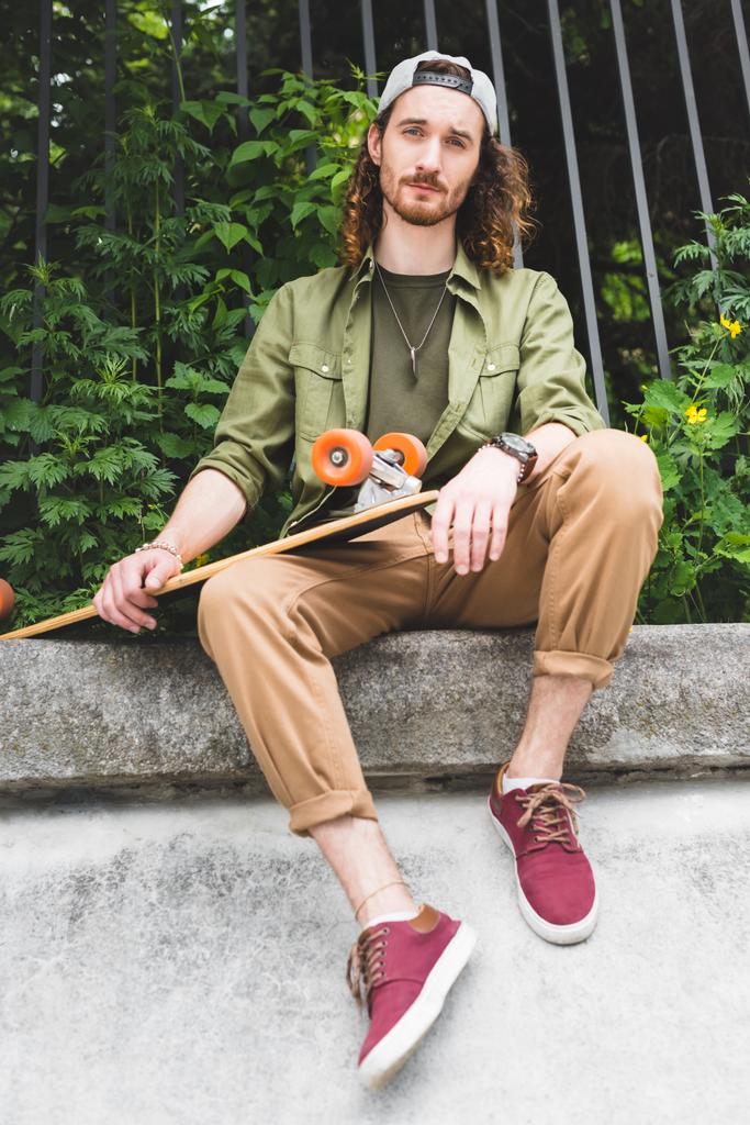 low angle view of handsome man holding skateboard in hands, looking at camera - Photo, Image