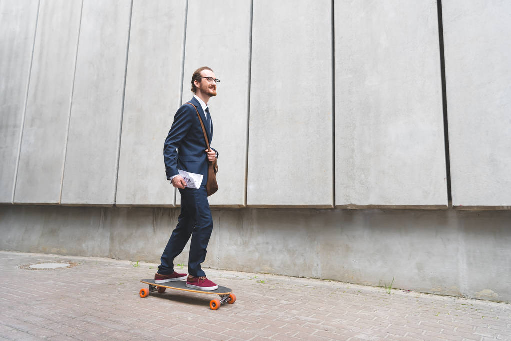 glücklicher Geschäftsmann in formeller Kleidung, der auf dem Skateboard fährt und wegschaut - Foto, Bild