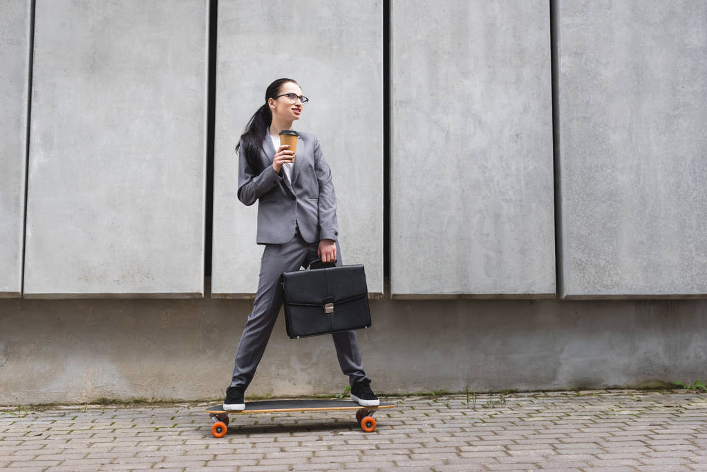 スケートボードに乗って、紙コップとブリーフケースを手に持つフォーマルな服装で陽気なビジネスウーマン - 写真・画像