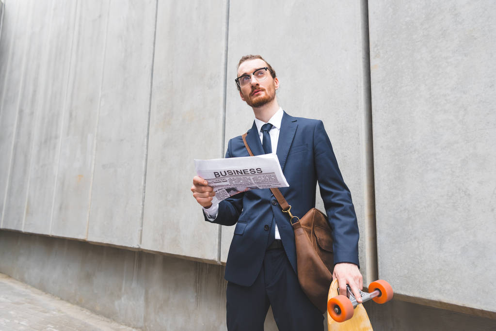низький кут зору бізнесмена в офіційному одязі, що стоїть біля скейтборду, тримаючи газету в руці
 - Фото, зображення
