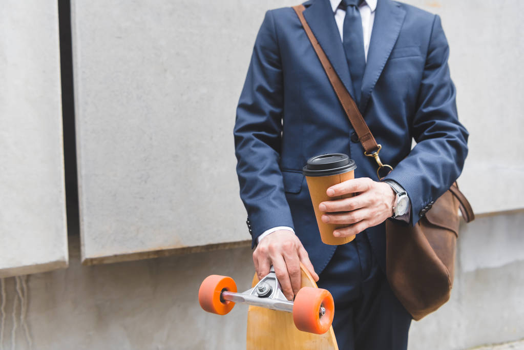 vista recortada de hombre de negocios en ropa formal de pie con monopatín y taza de papel en las manos
 - Foto, Imagen