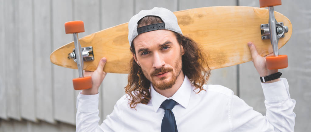 panoramic view of handsome businessman standing with skateboard, looking at camera - Photo, Image