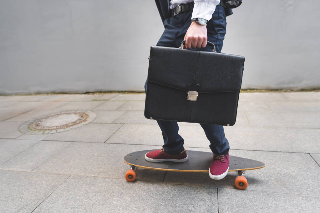 Ausgeschnittene Ansicht von Geschäftsmann mit Aktentasche in der Hand, der auf Skateboard fährt - Foto, Bild