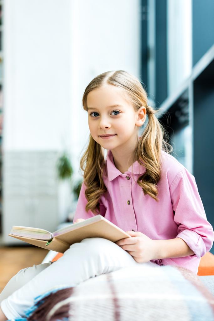 selective focus of cute and cheerful kid sitting and holding book in library  - Photo, Image