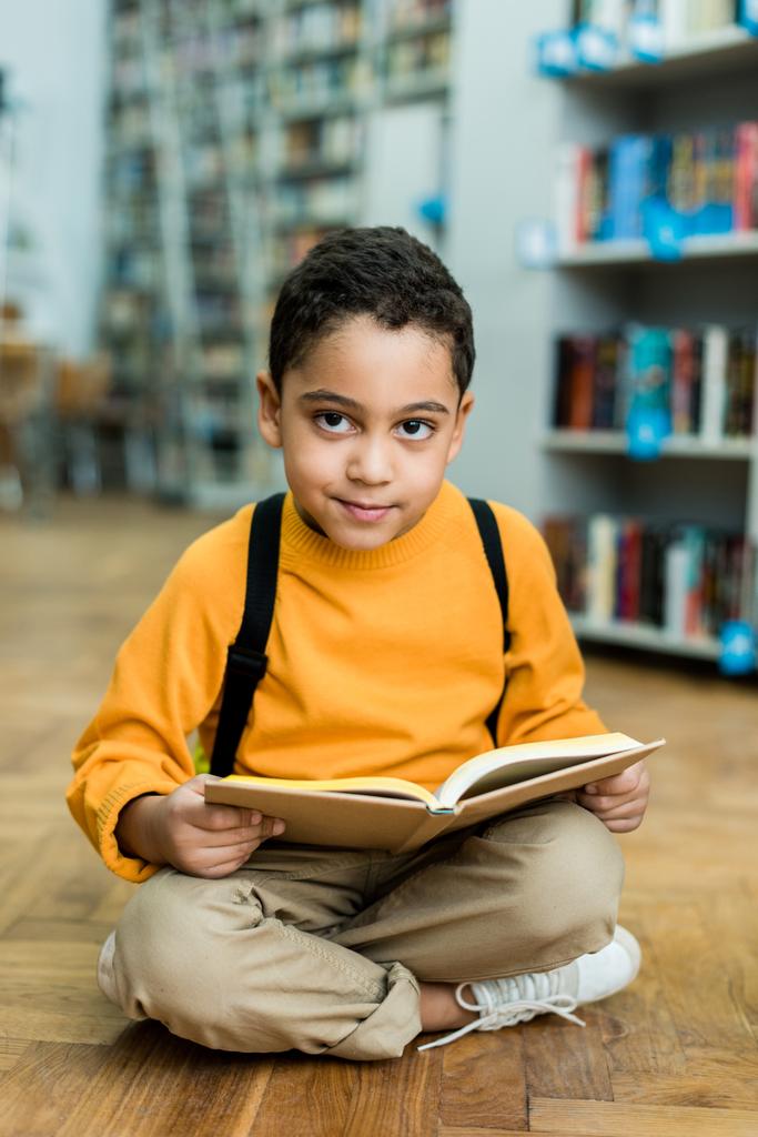 陽気なアフリカ系アメリカ人の少年が床に座って本を持っている  - 写真・画像