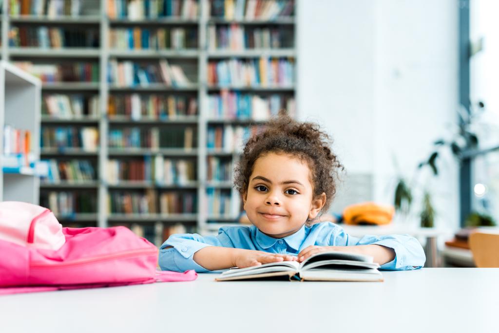 heureux afro-américain enfant assis et regardant la caméra près du livre et sac à dos rose
  - Photo, image