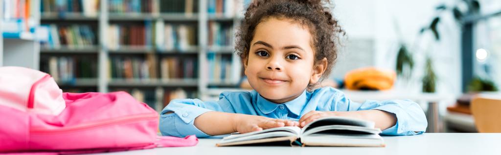 plano panorámico de niño afroamericano feliz sentado cerca del libro en la biblioteca
  - Foto, imagen