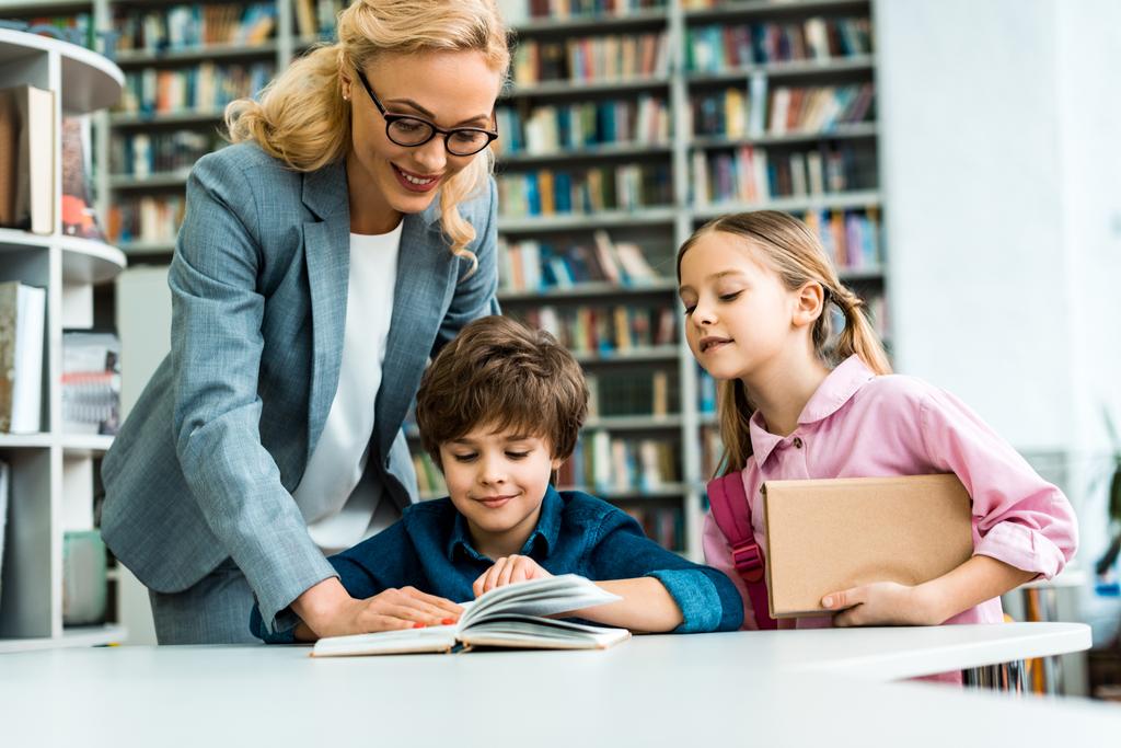веселый учитель в очках, стоящий рядом с милыми детьми, читающими книгу в библиотеке
  - Фото, изображение