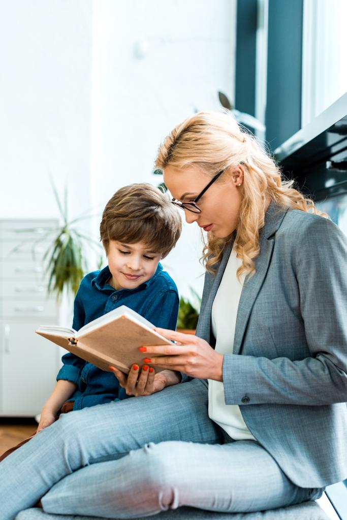 attractive woman in glasses sitting on floor and reading book with cute kid  - Photo, Image