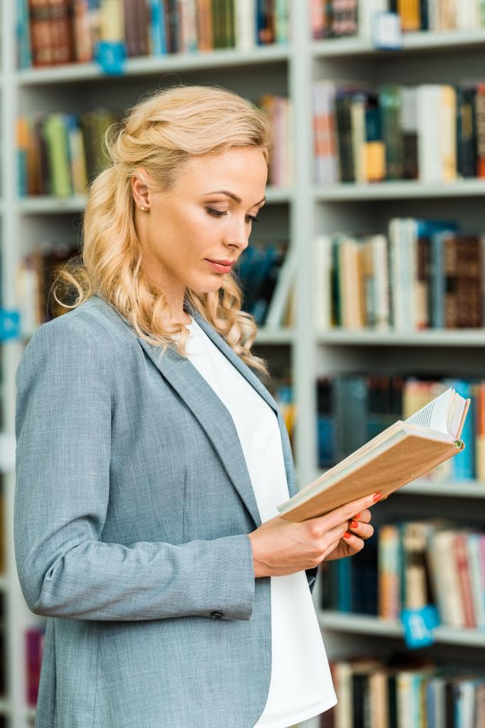 Piękne blond kobieta stoi i czytania książki w bibliotece  - Zdjęcie, obraz
