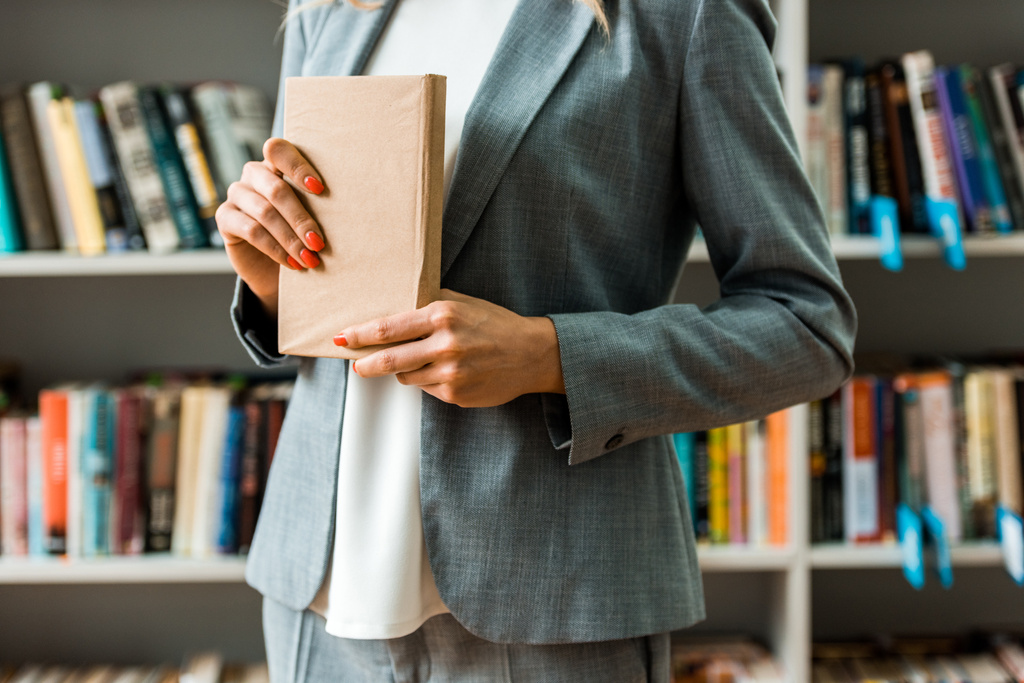 περικομμένη όψη της γυναίκας που στέκεται στη βιβλιοθήκη και κρατώντας το βιβλίο  - Φωτογραφία, εικόνα