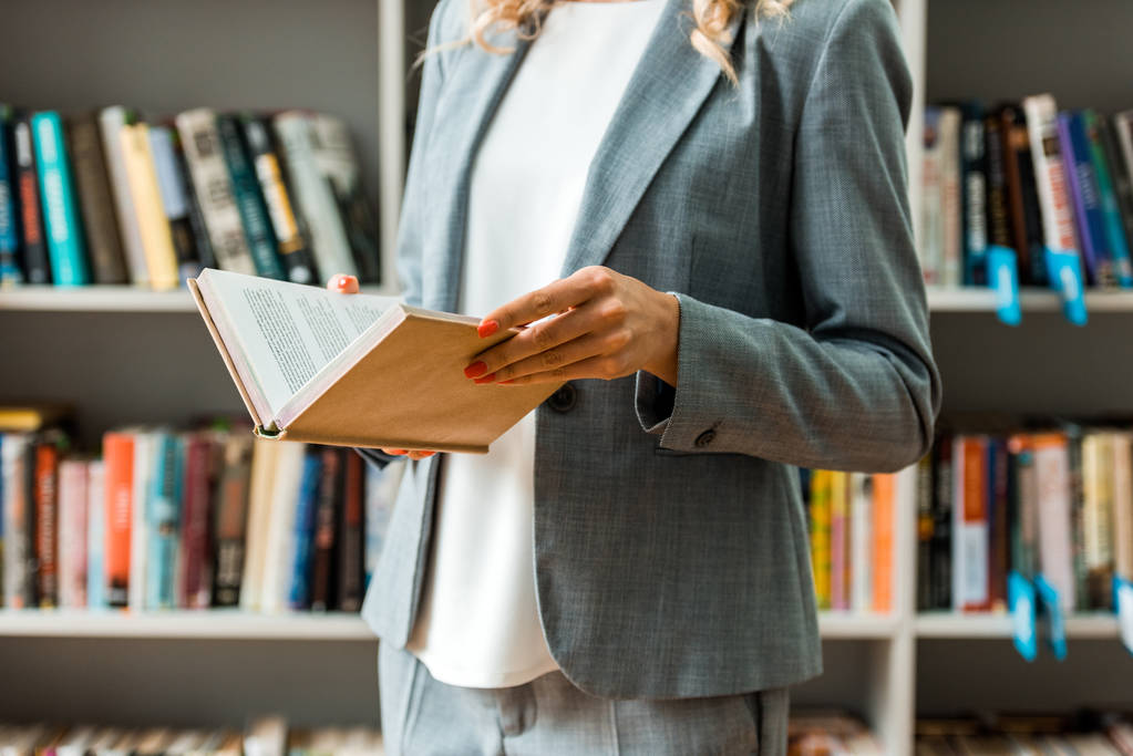 περικομμένη θέα της γυναίκας που κρατά το βιβλίο ενώ στέκεται στη βιβλιοθήκη  - Φωτογραφία, εικόνα