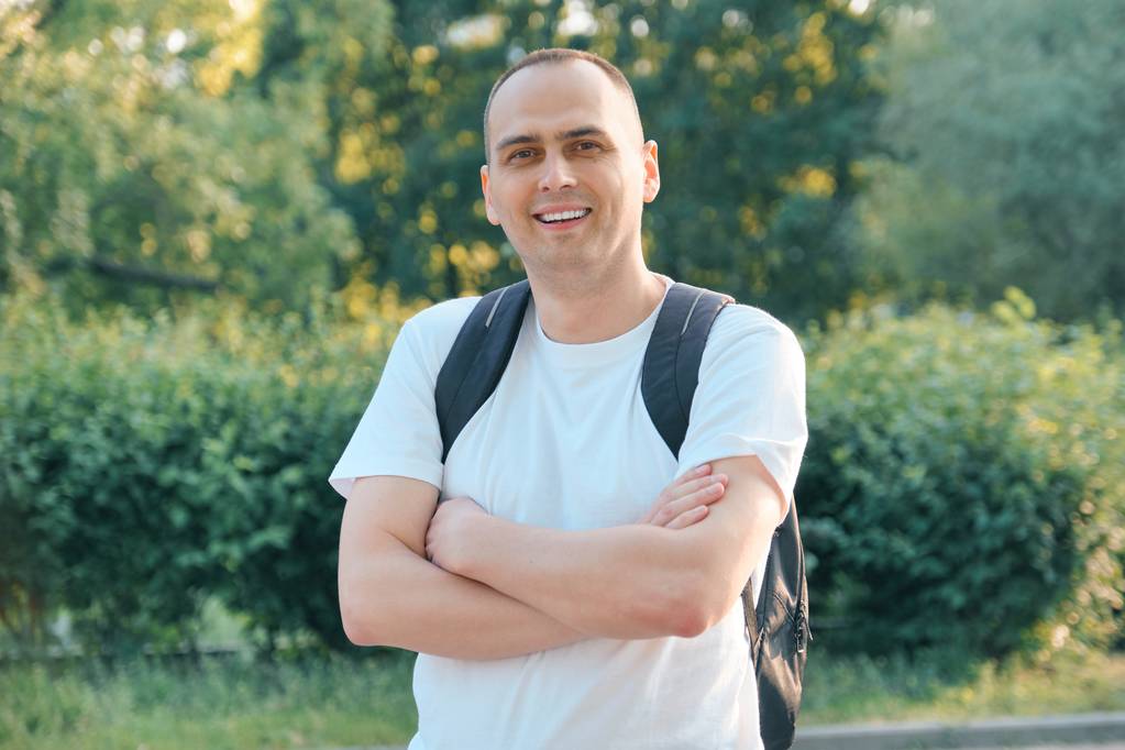 Outdoor portrait of mature smiling positive man. Confident male in white T-shirt with backpack with folded hands looking at the camera - Photo, Image