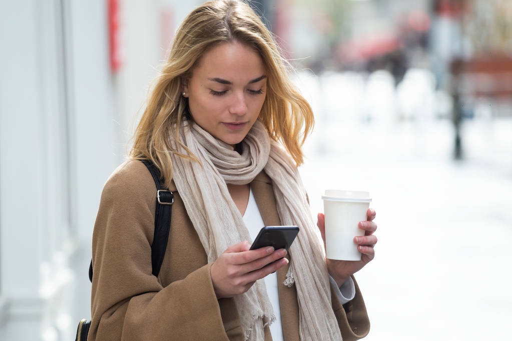 Mooie jonge vrouw met behulp van haar mobiele telefoon terwijl het houden van kopje koffie in de straat. - Foto, afbeelding