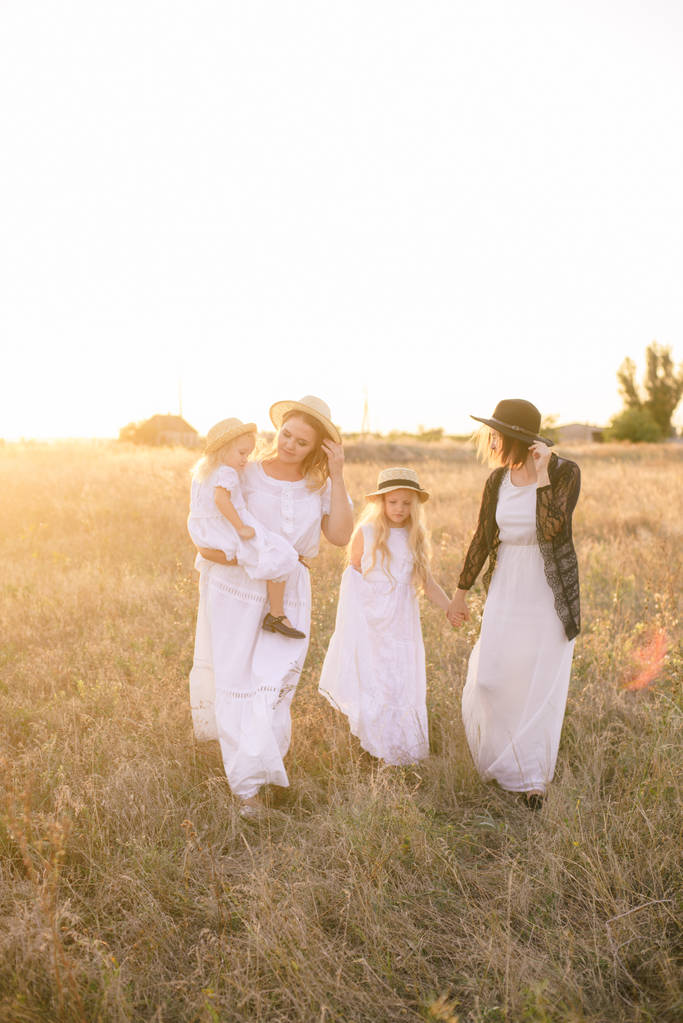 A young mother with her daughters and an aunt with blond hair in white dresses at sunset in the summer in a field of countryside. Several girls from the same family in the summer in the field - Photo, Image