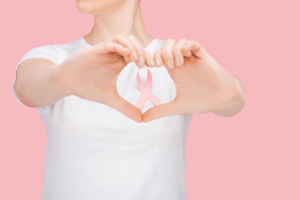 částečný pohled na ženu v bílém tričku, to je znamení srdce s rukama kolem růžové stuhy na rakovinu prsu, izolované na růžovém - Fotografie, Obrázek