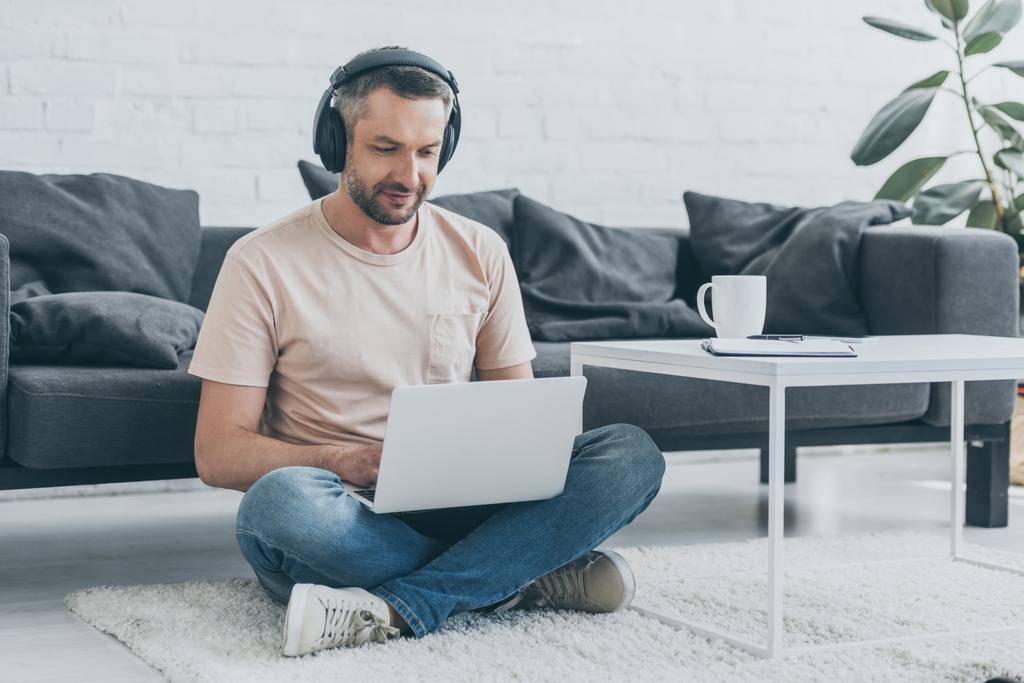 handsome man in headphones sitting on floor with crossed legs and using laptop - Photo, Image
