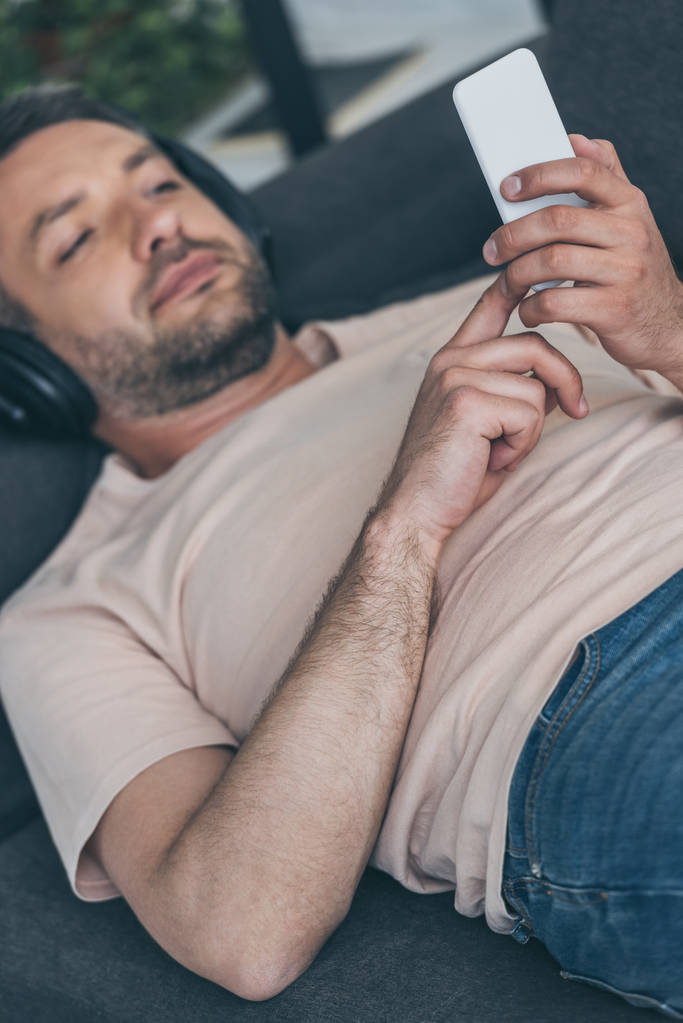 hombre guapo en los auriculares acostado en el sofá y el uso de teléfono inteligente
 - Foto, imagen
