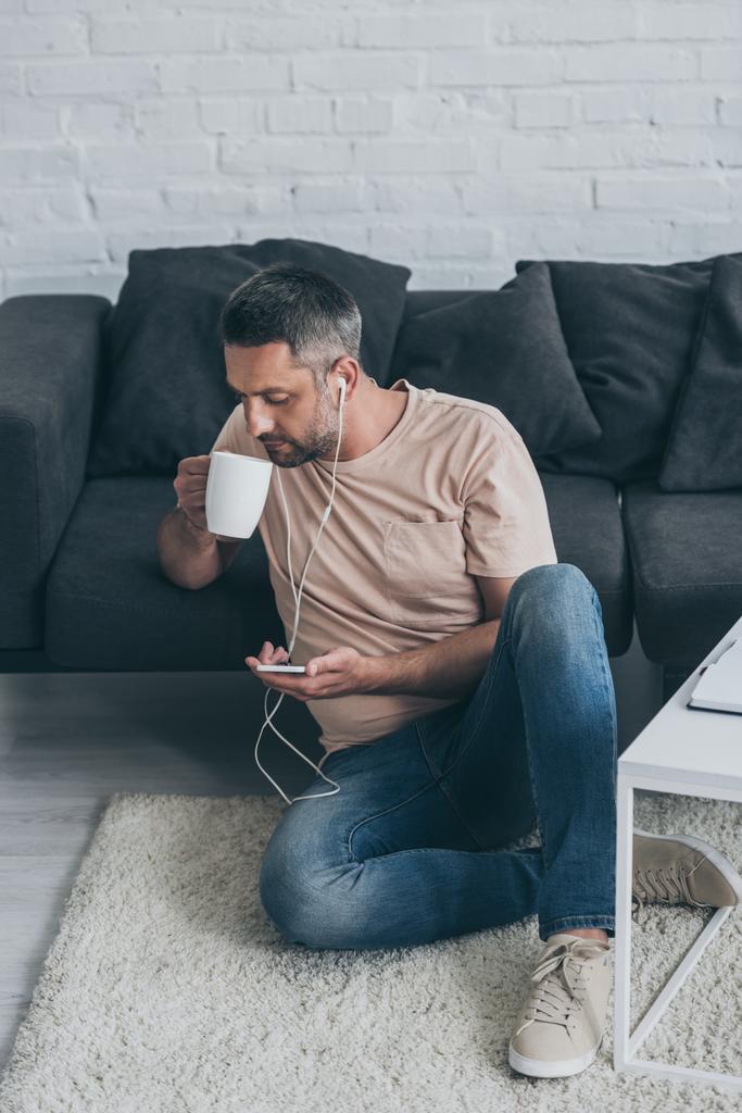 adult man sitting on floor, listening music in earphones, using smartphone and drinking coffee - Photo, Image