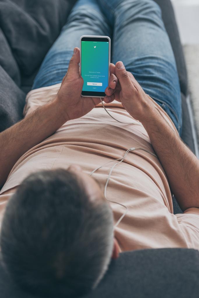 キエフ、 ウクライナ - 2019年5月5日:ソファに横たわり、画面上のツイッターアプリでスマートフォンを保持している男性の選択的な焦点. - 写真・画像