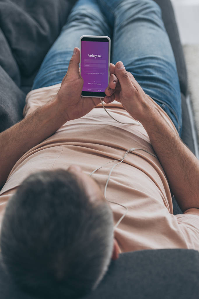 KYIV, UCRANIA - 5 de mayo de 2019: Enfoque selectivo del hombre en los auriculares acostado en el sofá y sosteniendo el teléfono inteligente con la aplicación de Instagram en la pantalla
. - Foto, Imagen