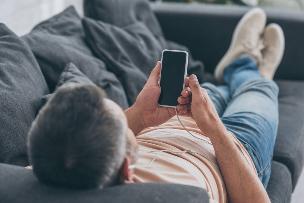 ソファに横たわり、空白の画面でスマートフォンを保持するイヤホンの男性の選択的な焦点 - 写真・画像