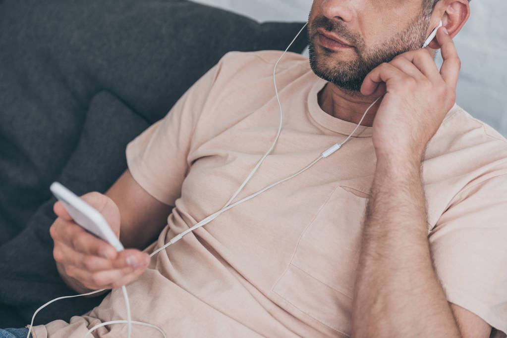 cropped view of adult man holding smartphone and listening music in earphones - Photo, Image