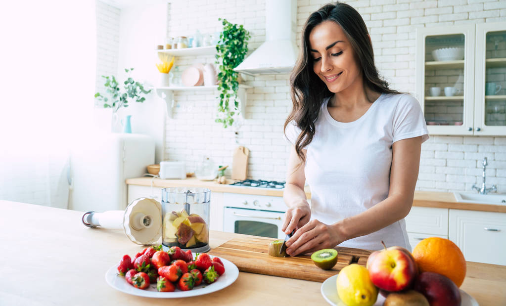 Beautiful woman making fruits smoothies with blender. Healthy eating lifestyle concept portrait of beautiful young woman preparing drink with bananas, strawberry and kiwi at home in kitchen. - Photo, Image