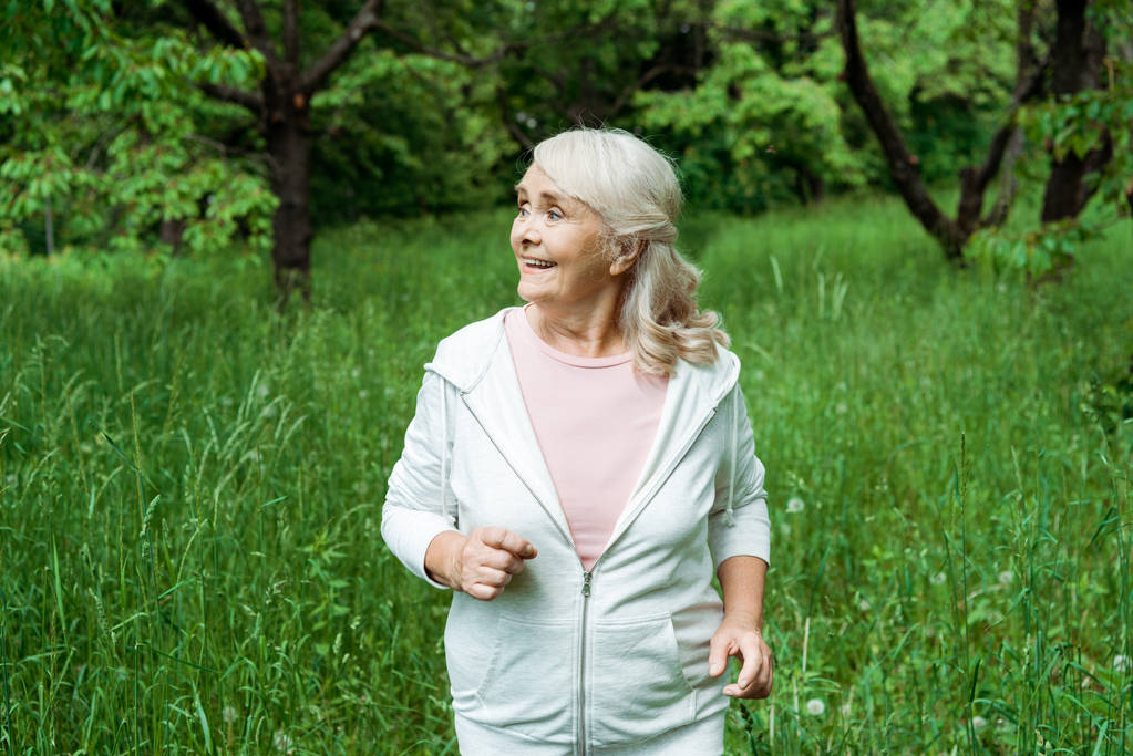 Szczęśliwa Starsza kobieta z szarymi włosami działa w zielonym parku  - Zdjęcie, obraz