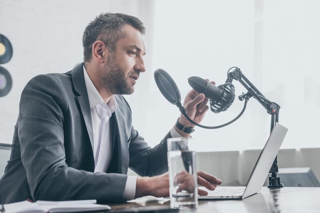 komea radio isäntä puhuu mikrofonilla istuessaan työpaikalla lähellä kannettavaa tietokonetta ja lasi vettä
 - Valokuva, kuva