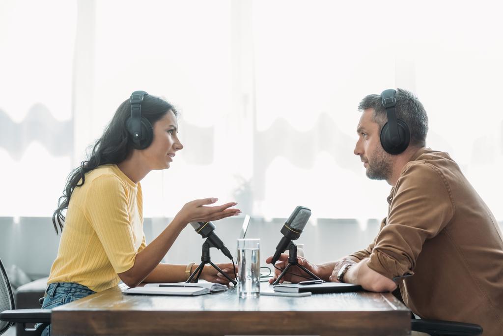 twee serieuze radio-hosts in hoofdtelefoons praten terwijl zittend op werkplek in Studio - Foto, afbeelding