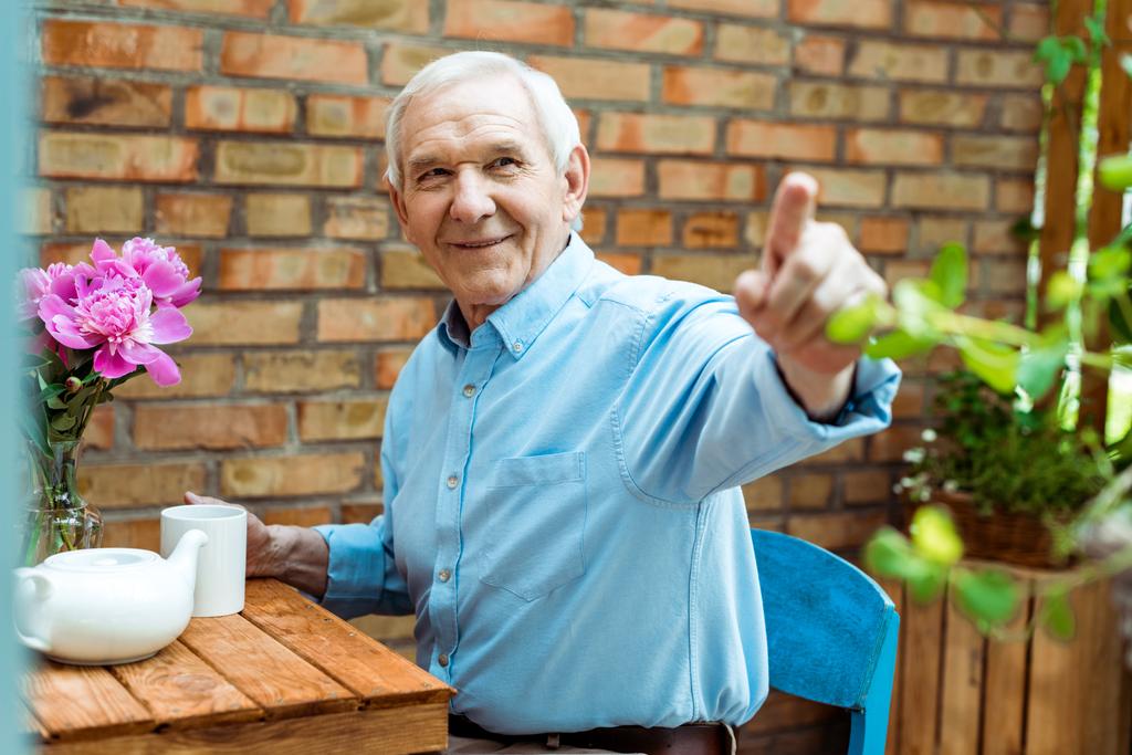 Selektiver Fokus eines glücklichen Seniors, der mit dem Finger zeigt, während er Tasse hält  - Foto, Bild