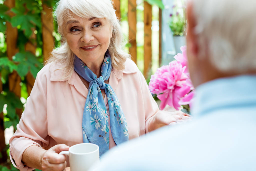 Selektiver Fokus einer glücklichen Seniorin, die ihren Mann ansieht, während sie eine Tasse hält  - Foto, Bild