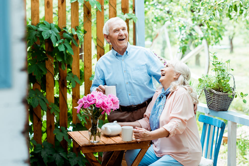 ピンクの花の近くで陽気な妻と笑う幸せな先輩男性の選択的な焦点  - 写真・画像