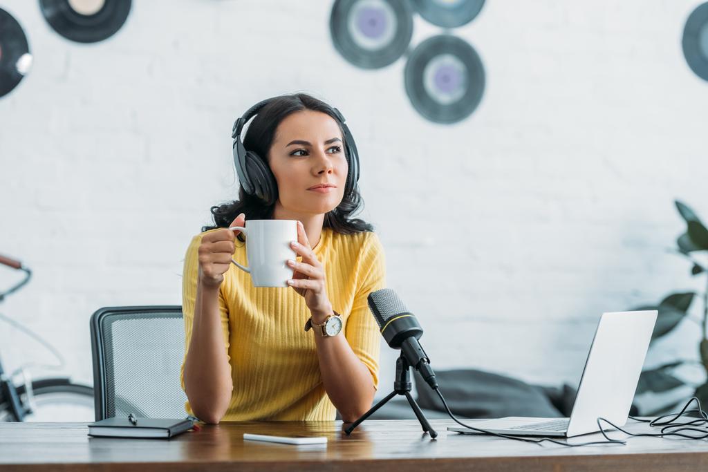 hôte radio coûteux regardant loin tout en étant assis sur le lieu de travail et tenant tasse de café
 - Photo, image