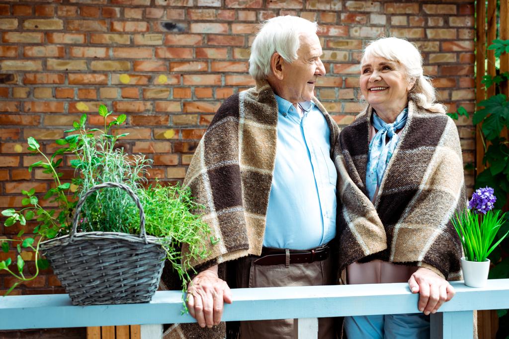 花鉢の近くに格子状の毛布の中に立ちながら微笑む幸せな年金生活者  - 写真・画像