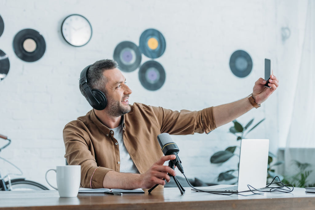 Veselý rozhlasový hostitel ve sluchátkách s smartphením při sezení na pracovišti  - Fotografie, Obrázek