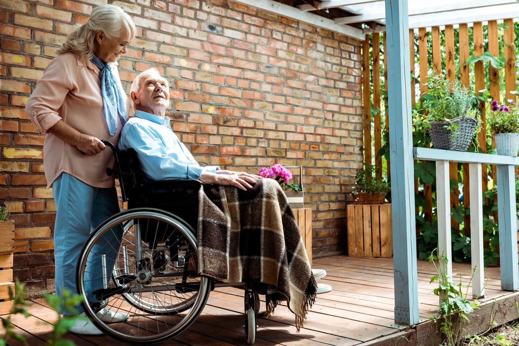 valikoiva painopiste iloinen vanhempi nainen seisoo lähellä vammainen aviomies pyörätuolissa
  - Valokuva, kuva