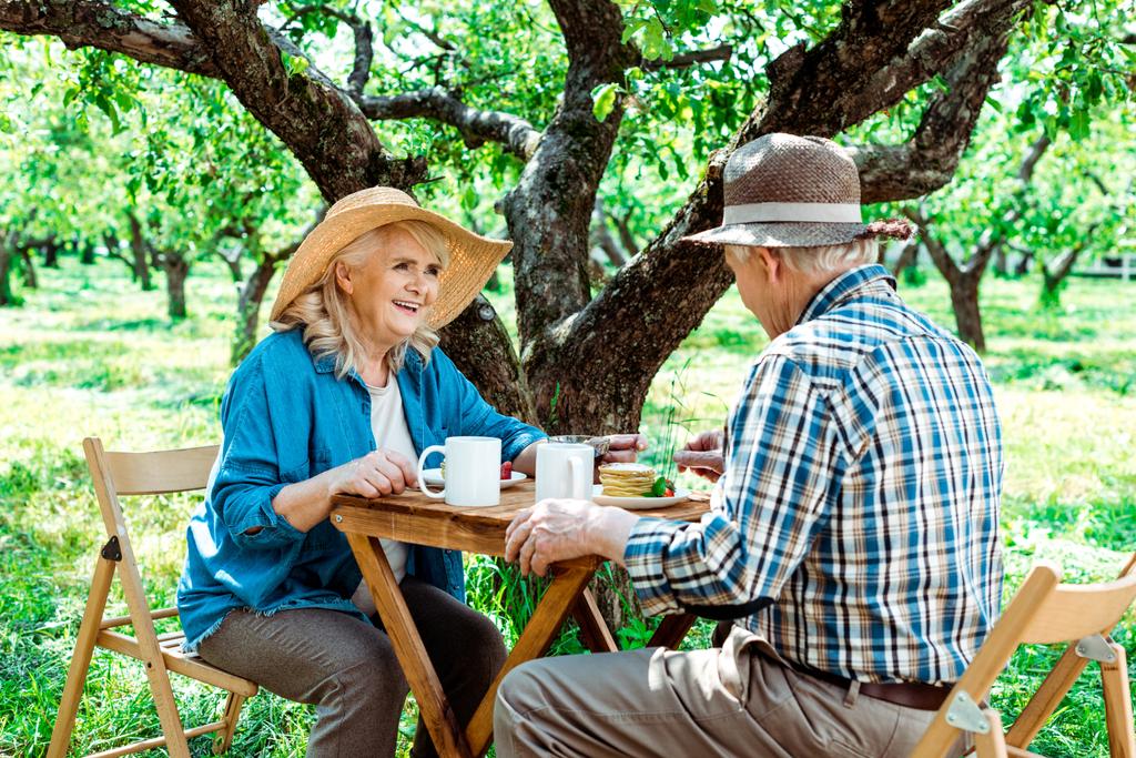 весела старша жінка в солом'яному капелюсі дивиться на чоловіка біля дерева
  - Фото, зображення