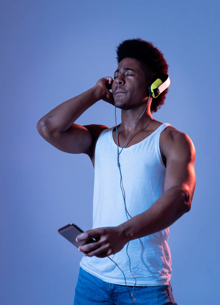 Fuerte afroamericano joven escuchando música en la aplicación de teléfonos inteligentes en busca de la mejor canción en línea con auriculares cantando y bailando. Retrato con pista de baile disco luz en Música y tecnología
. - Foto, Imagen
