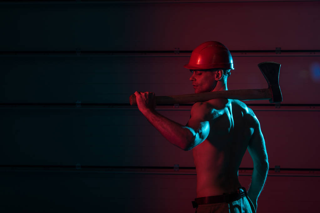 Sexy γυμνός πυροσβέστης σε προστατευτικό κράνος κρατώντας επίπεδη κεφάλι τσεκούρι στο σκοτάδι - Φωτογραφία, εικόνα