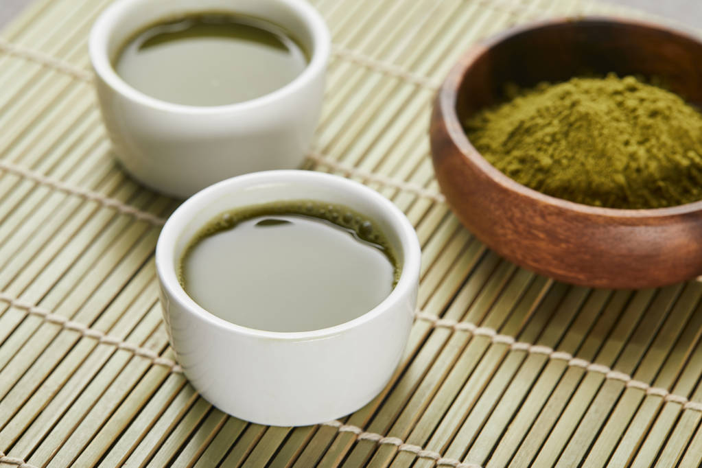 selektivní zaměření bílých šálků s čajem v blízkosti zeleného prášku matča v dřevěné misce - Fotografie, Obrázek