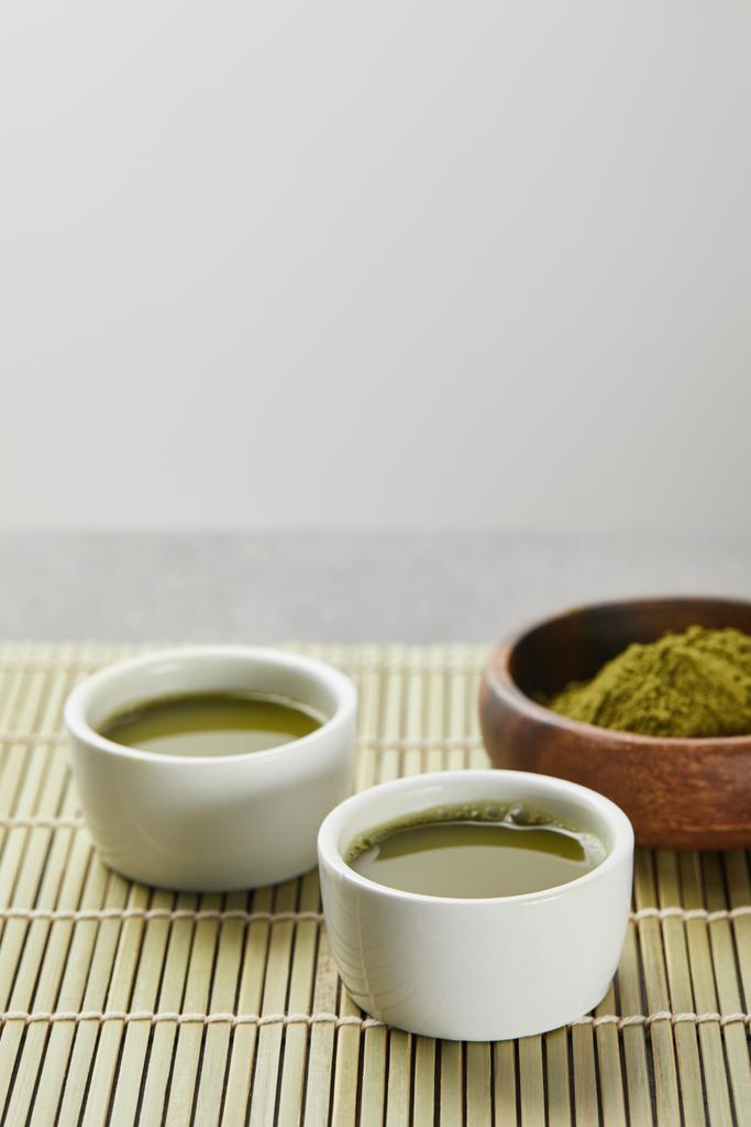 selektivní zaměření zeleného prášku na matča v dřevěné misce blízko bílých pohárků s čajem na bambusovou podložku - Fotografie, Obrázek