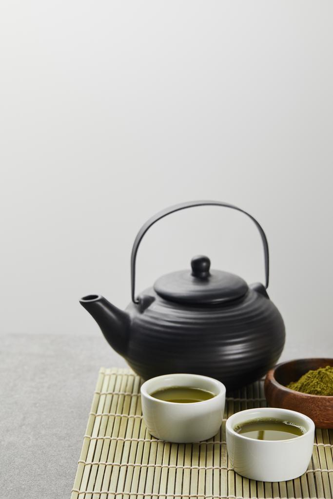 επιλεκτική εστίαση της πράσινης σκόνης μάτσα σε ξύλινο μπολ κοντά σε μαύρη τσαγιέρα και φλιτζάνια με τσάι σε μπαμπού τραπέζι χαλάκι - Φωτογραφία, εικόνα