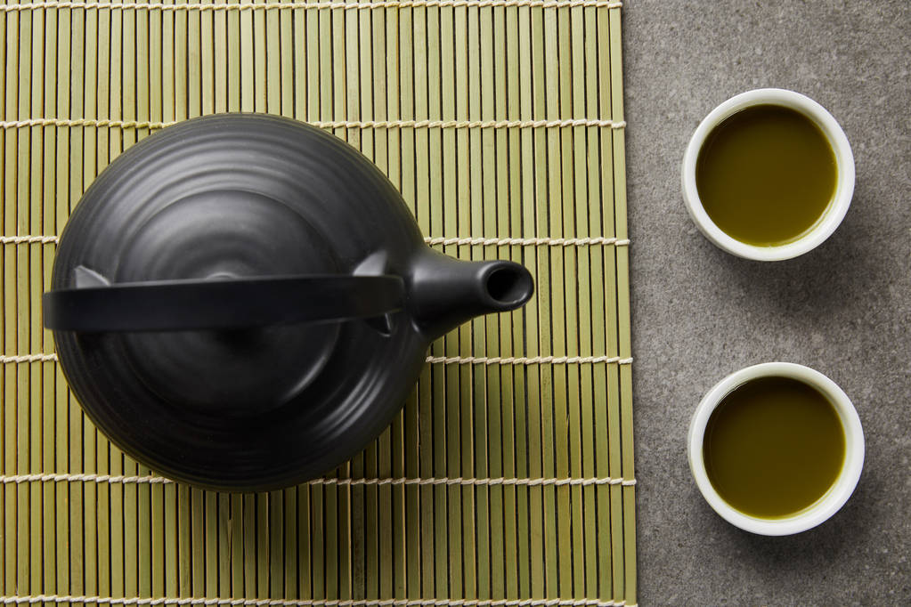 Top uitzicht van witte kopjes met groene Matcha thee in de buurt van zwarte theepot op bamboe placemat - Foto, afbeelding