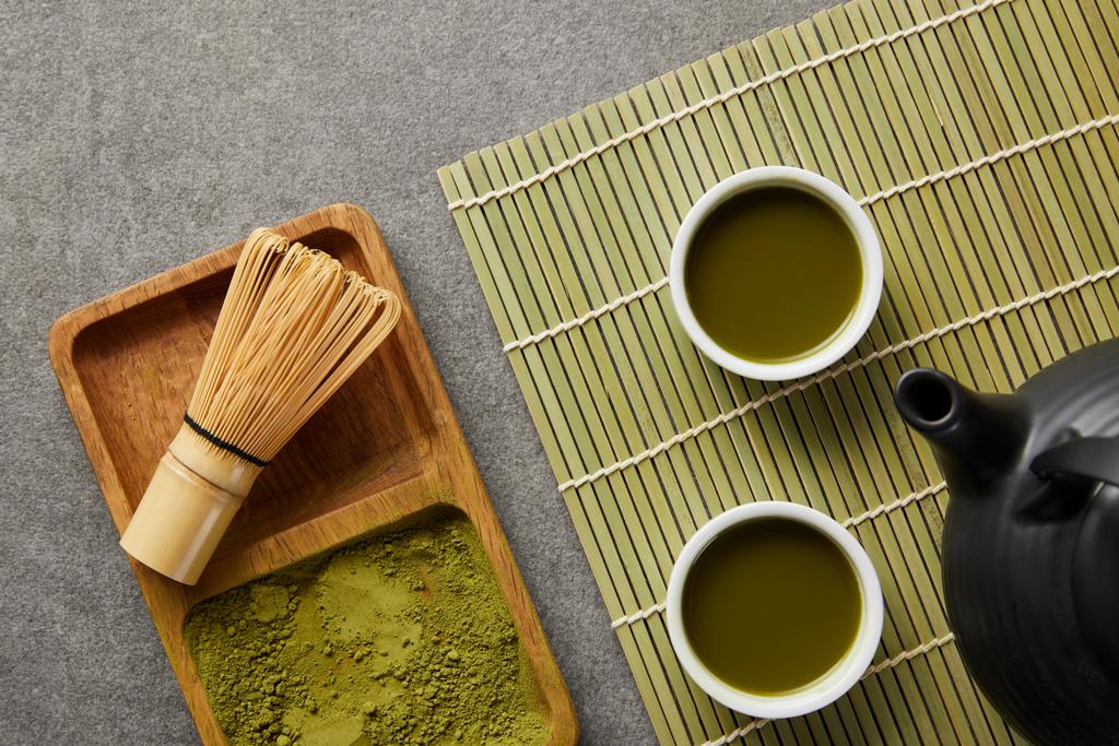 Widok z góry zielonego proszku Matcha z bambusa trzepaczka w drewnianej płycie w pobliżu czajniczek i białych filiżanek z zielonej herbaty na stole mat - Zdjęcie, obraz